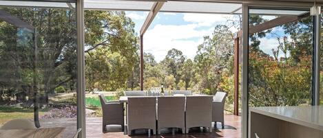 A modern outdoor dining area with a glass-enclosed patio, wicker furniture, and a view of a natural landscape.