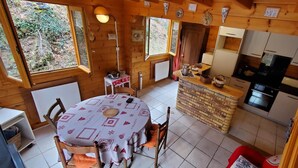 A wooden cabin with a round dining table, chairs, a kitchen area, and a view of the outdoors. - AU DEPART DE MULTIPLES RANDONNEES (Murol)