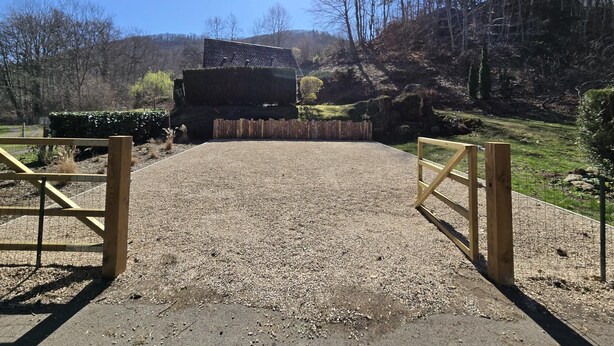 A gravel driveway leading to a wooden gate, surrounded by a fenced area with a building in the background. - AU DEPART DE MULTIPLES RANDONNEES (Murol)