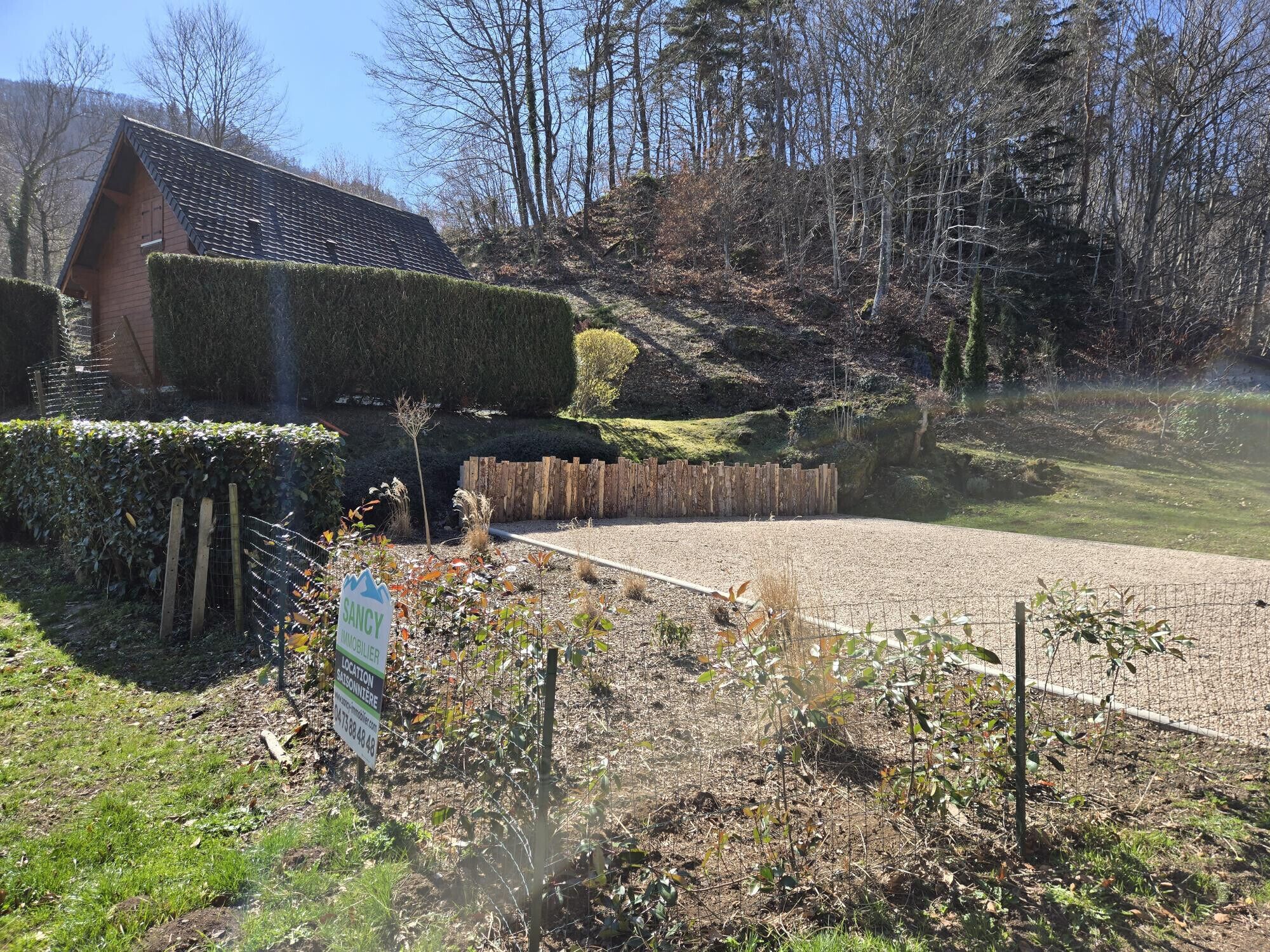 A rural property with a wooden house, a gravel driveway, and a fenced area with various plants.