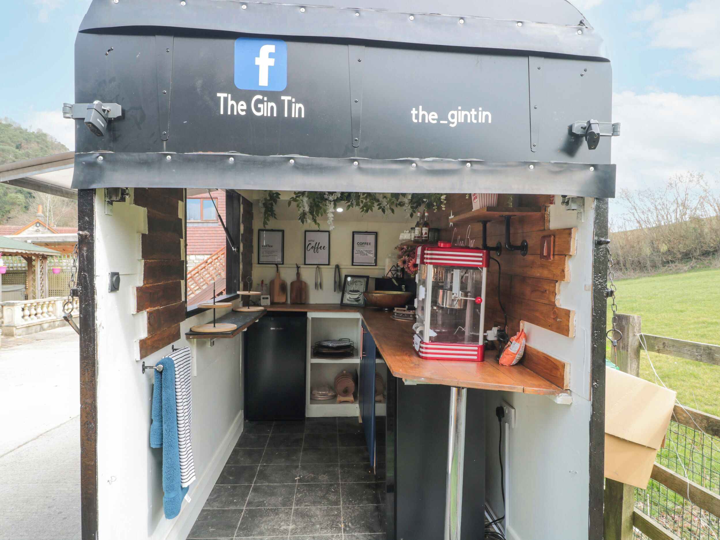 Una pequeña cafetería al aire libre llamada "The Gin Tin" con un interior rústico, una máquina de café y el logo de Facebook en el toldo.