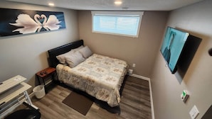 A bedroom with a bed, a desk, a chair, a TV, and a window.