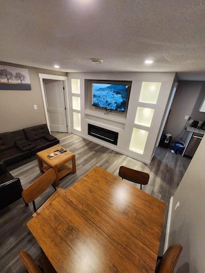 A modern living room with a wooden dining table, a dark-colored sofa, and a flat-screen TV mounted on the wall.