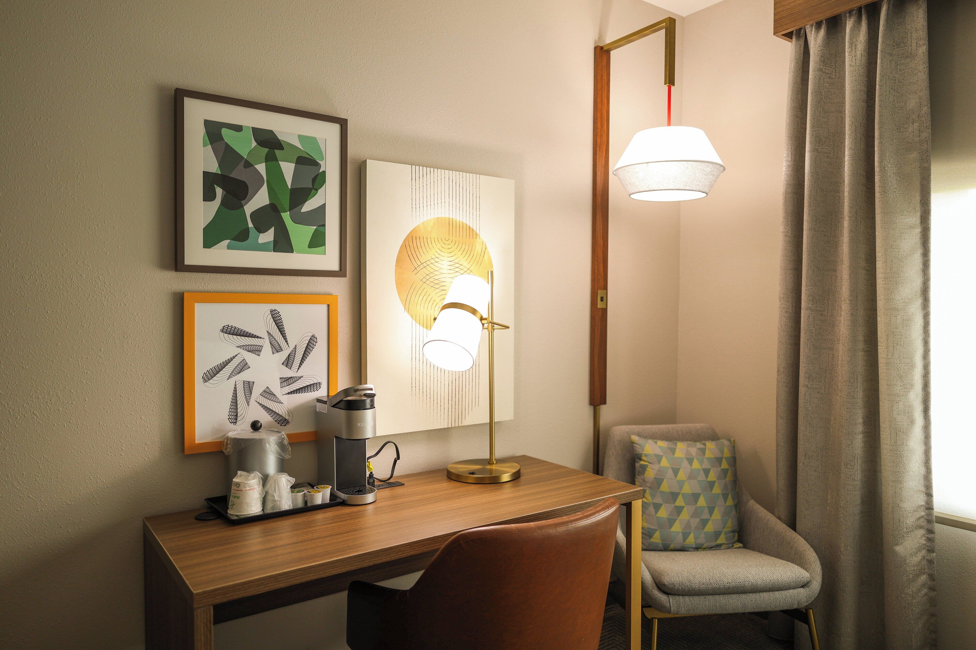 A hotel room with a wooden desk, a coffee maker, a lamp, and a chair.