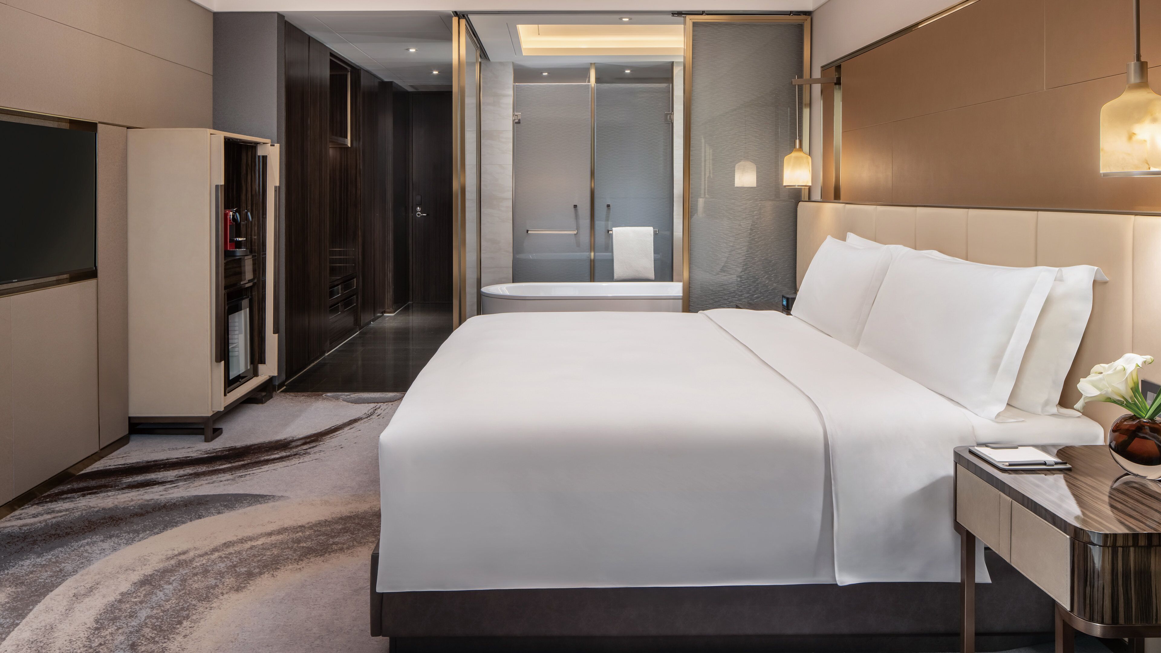 A modern hotel room with a large bed, a walk-in closet, a shower area, and a bedside table with a lamp.