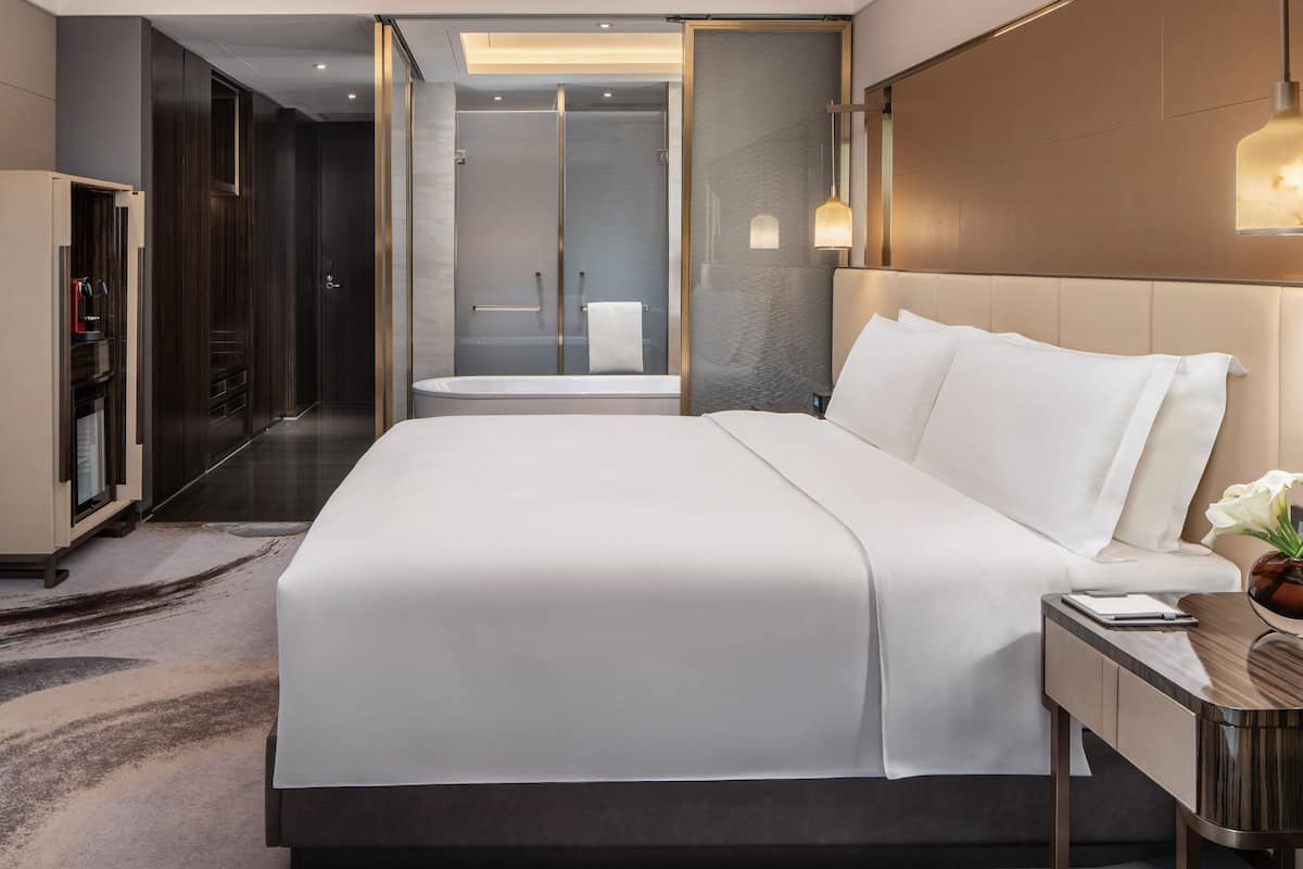 Une chambre d’hôtel moderne dotée d’un grand lit, d’un dressing, d’un espace douche et d’une table de chevet avec une lampe.