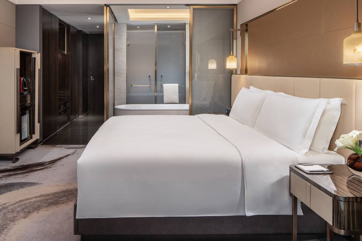 A modern hotel room with a large bed, a walk-in closet, a shower area, and a bedside table with a lamp.