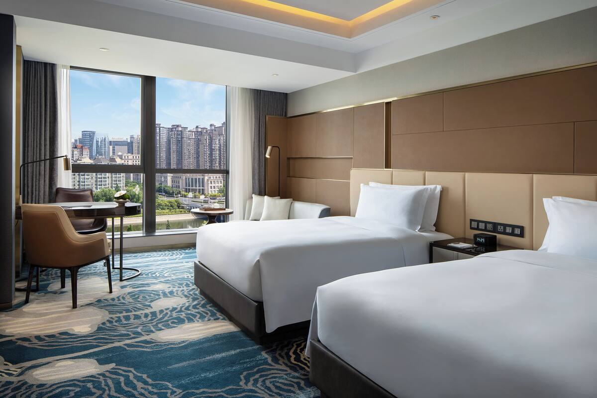 Une chambre d’hôtel avec deux lits, un bureau, une chaise, une grande fenêtre donnant sur la ville et un design moderne.