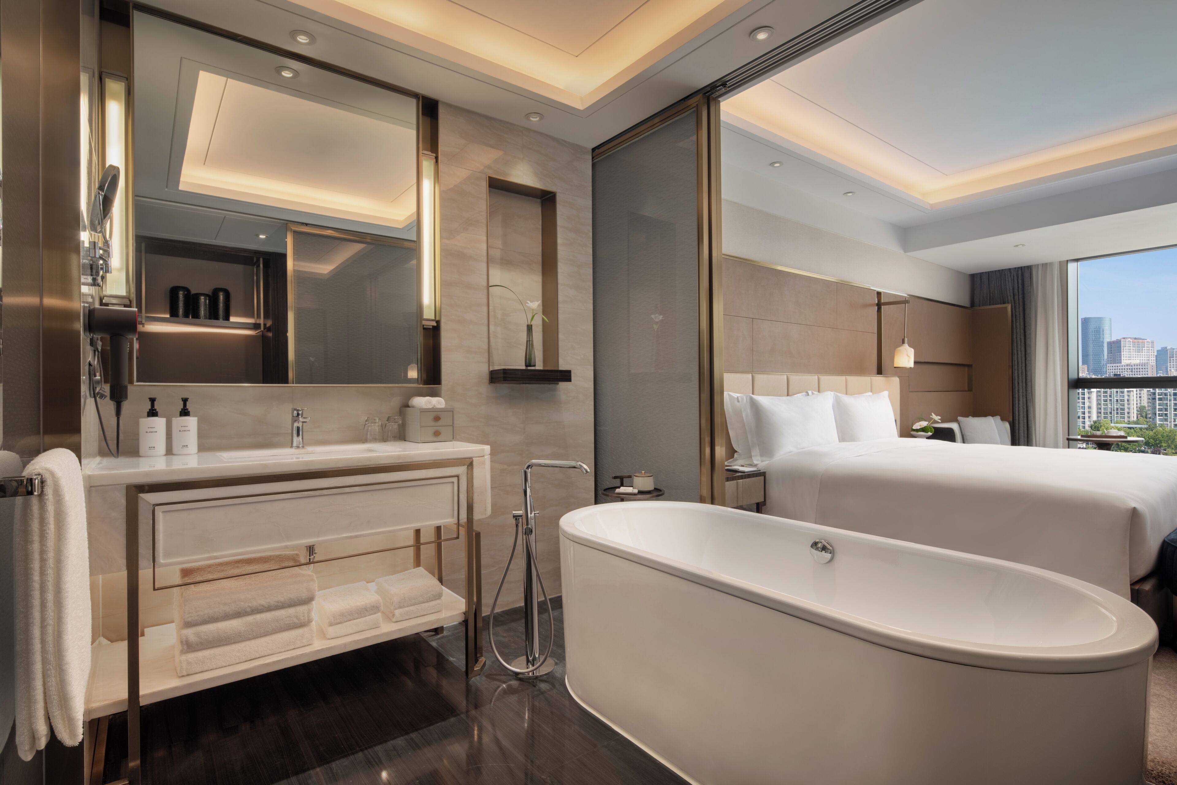 A modern hotel bathroom with a freestanding bathtub, a vanity with a mirror, and a large window offering a city view.