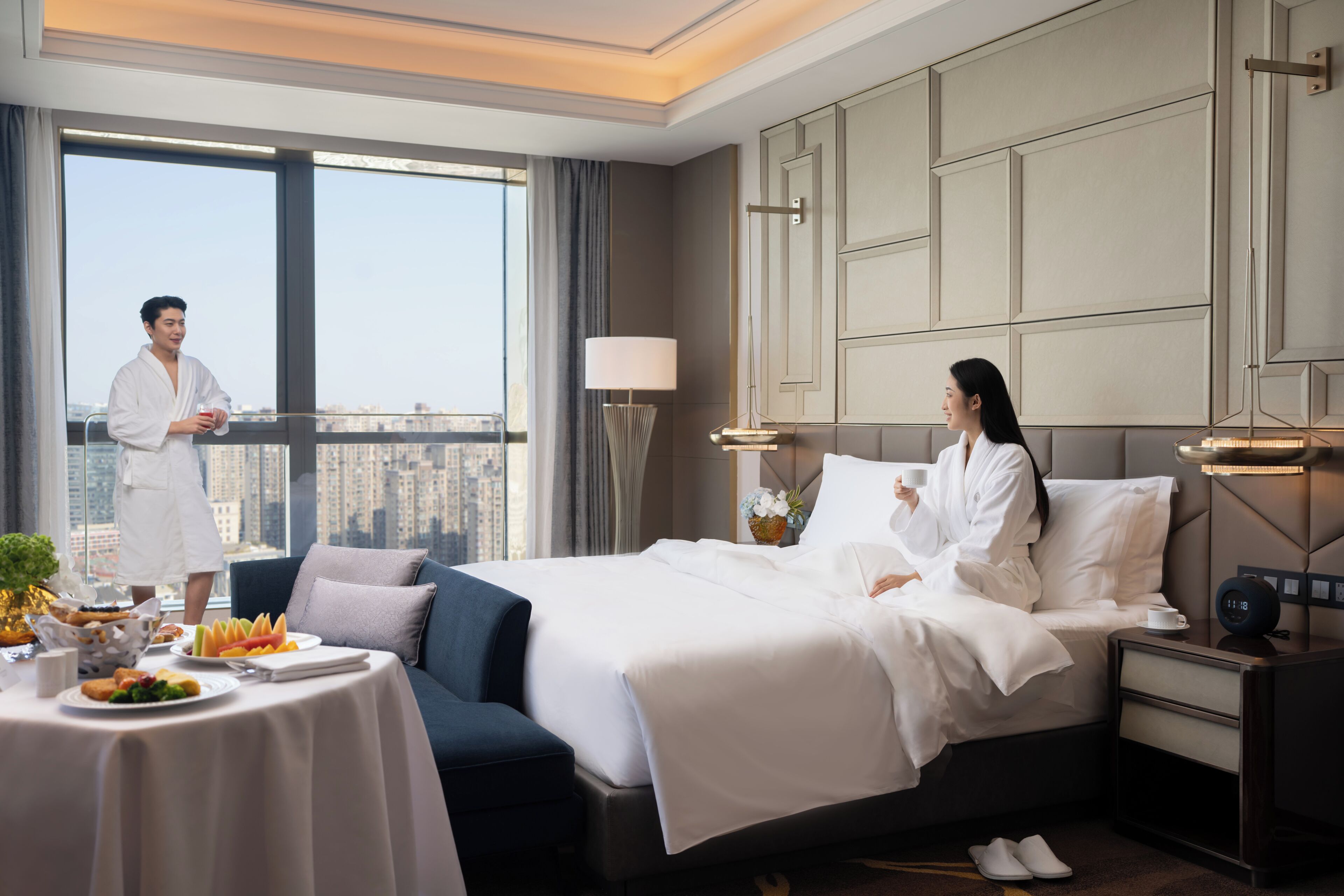 A hotel room with a large bed, a bedside table with a lamp, a nightstand with a clock, and a view of the city through the window.
