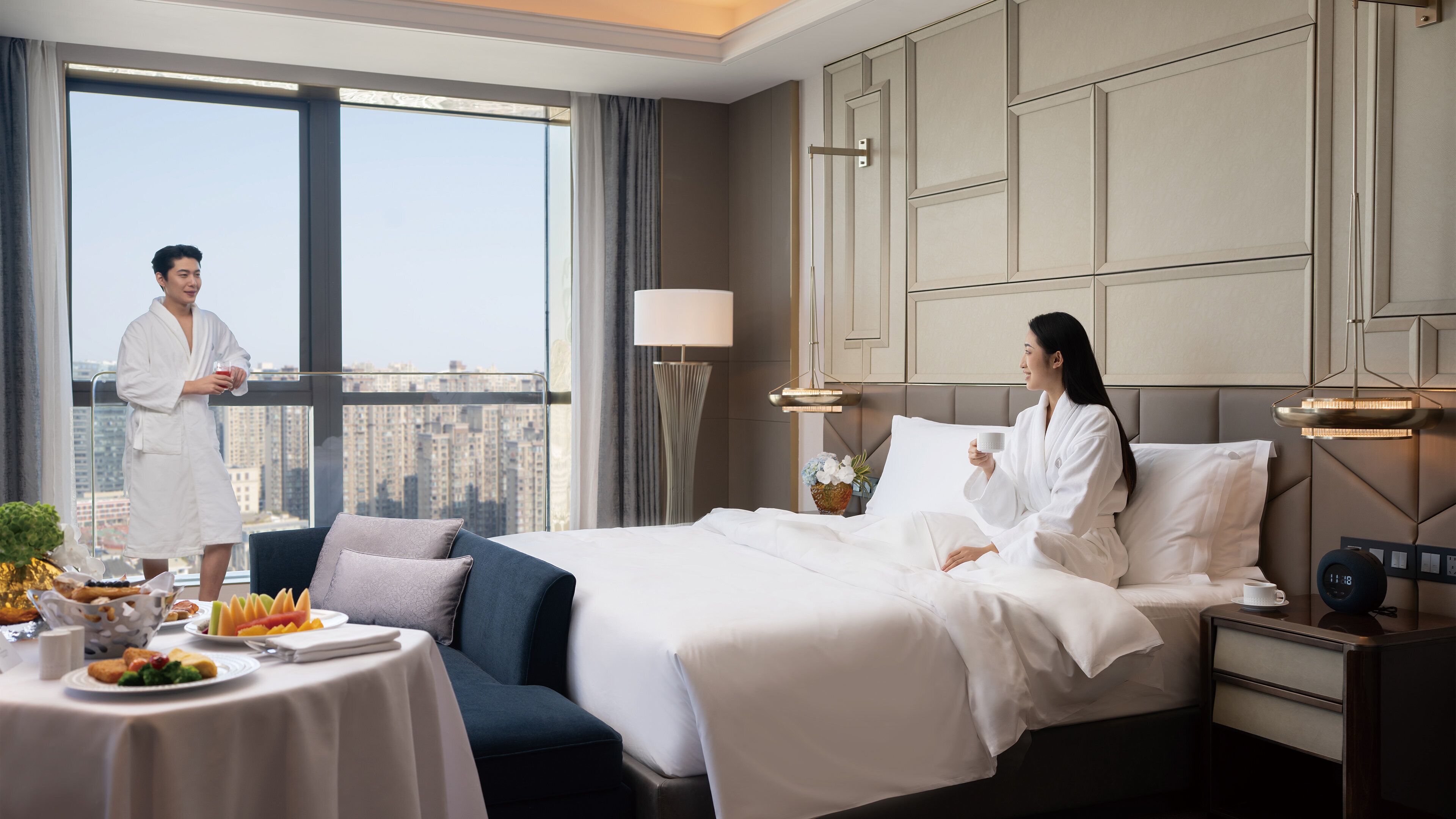 A hotel room with a large bed, a bedside table with a lamp, a nightstand with a clock, and a view of the city through large windows.