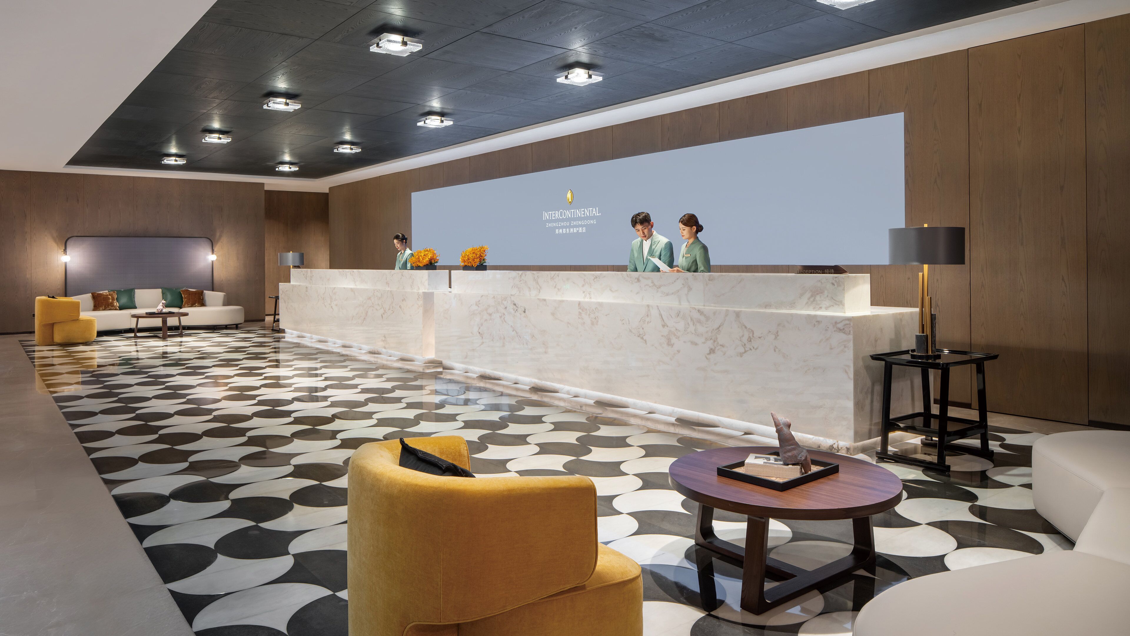 Uma área de recepção de hotel com um balcão de mármore, um sofá, uma poltrona amarela e um piso estampado.