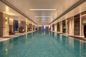 Uma piscina interior extensa com piso ladrilhado, rodeada por paredes com grandes janelas e luminárias modernas.