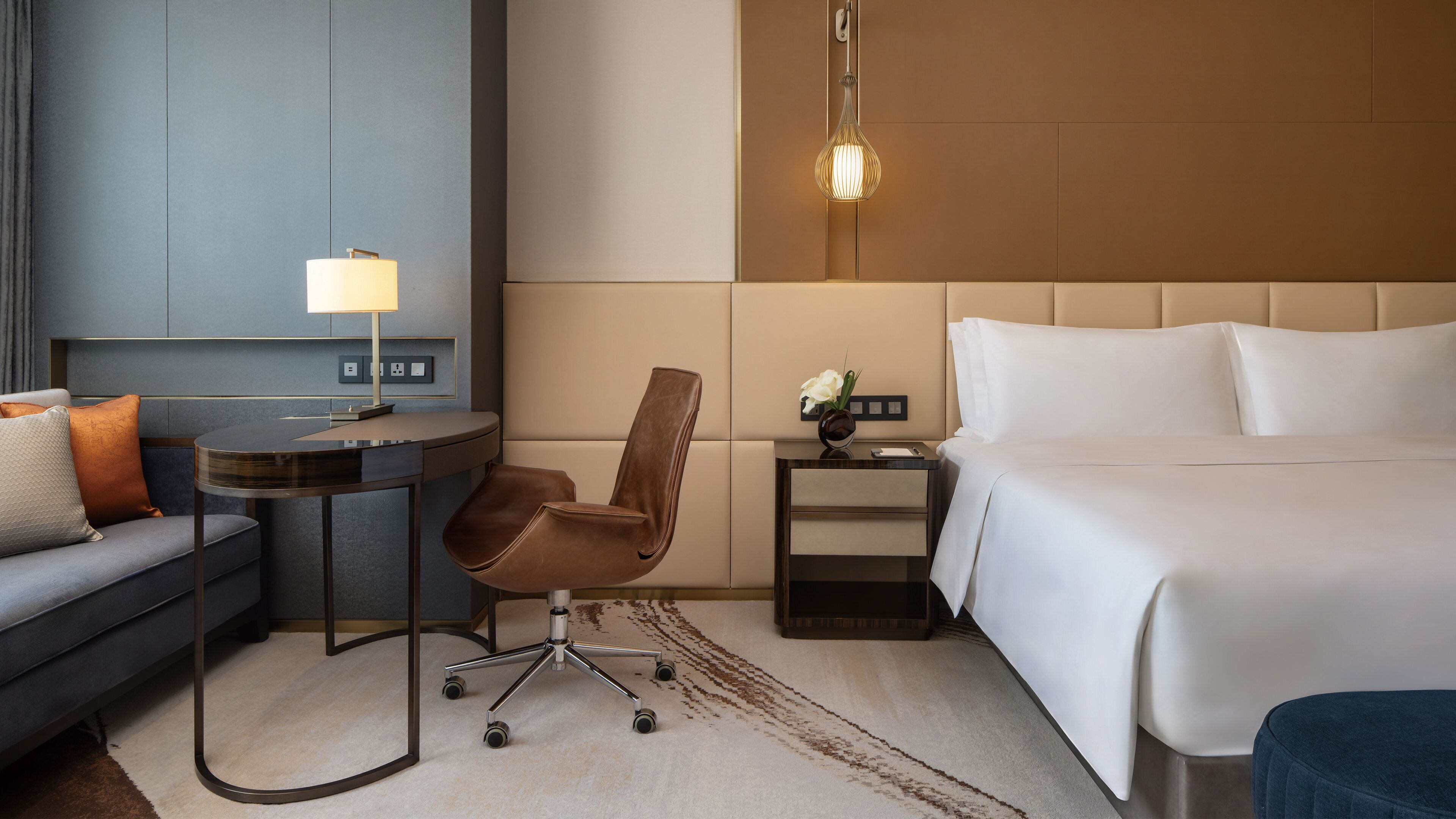 A hotel room with a bed, a desk, a chair, a lamp, and a nightstand with a vase of flowers.