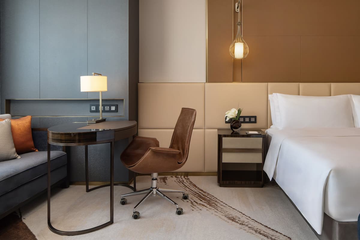 A hotel room with a bed, a desk, a chair, a lamp, and a nightstand with a vase of flowers.