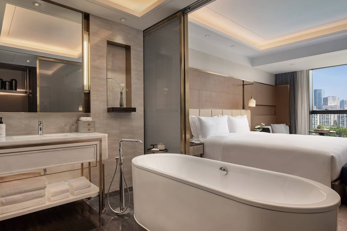 Une salle de bain d’hôtel moderne, dotée d’une grande baignoire, d’un meuble-lavabo avec miroir et d’un lit offrant une vue sur la ville.