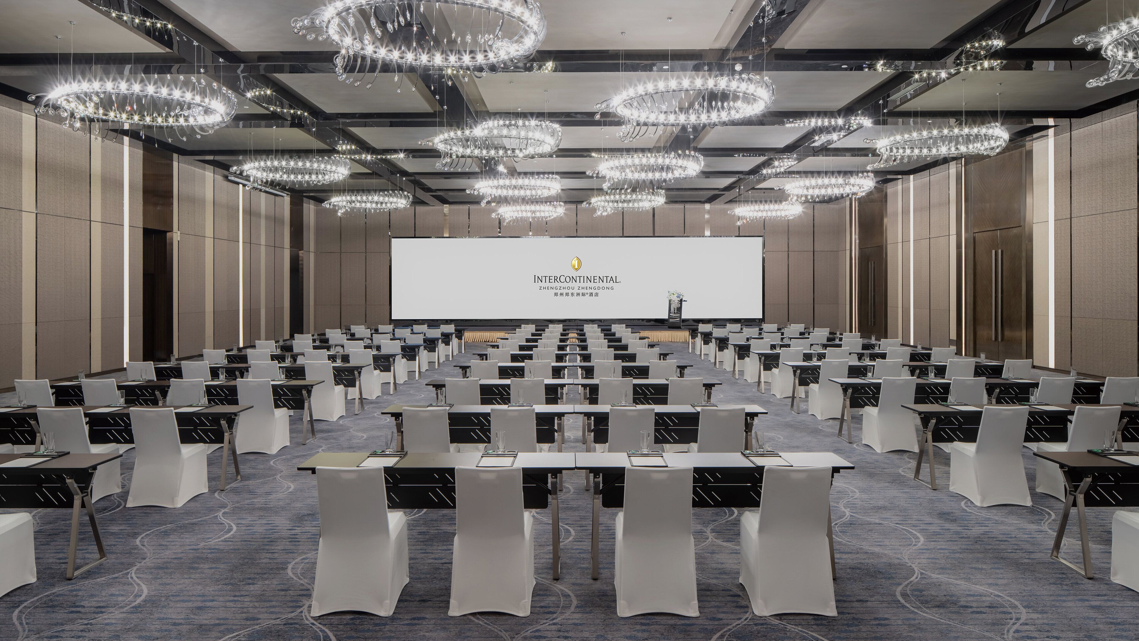 A conference room with rows of tables and chairs, a large screen at the front, and multiple chandeliers.