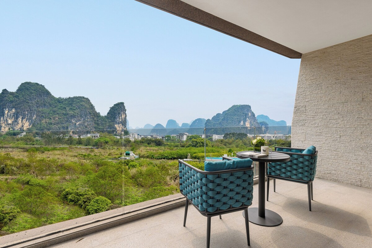 Un balcon avec deux chaises et une table, donnant sur un paysage montagneux verdoyant, avec des bâtiments visibles au loin.