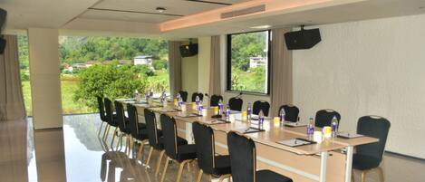 A conference room with a long table, chairs, and a large window offering a view of greenery and buildings.