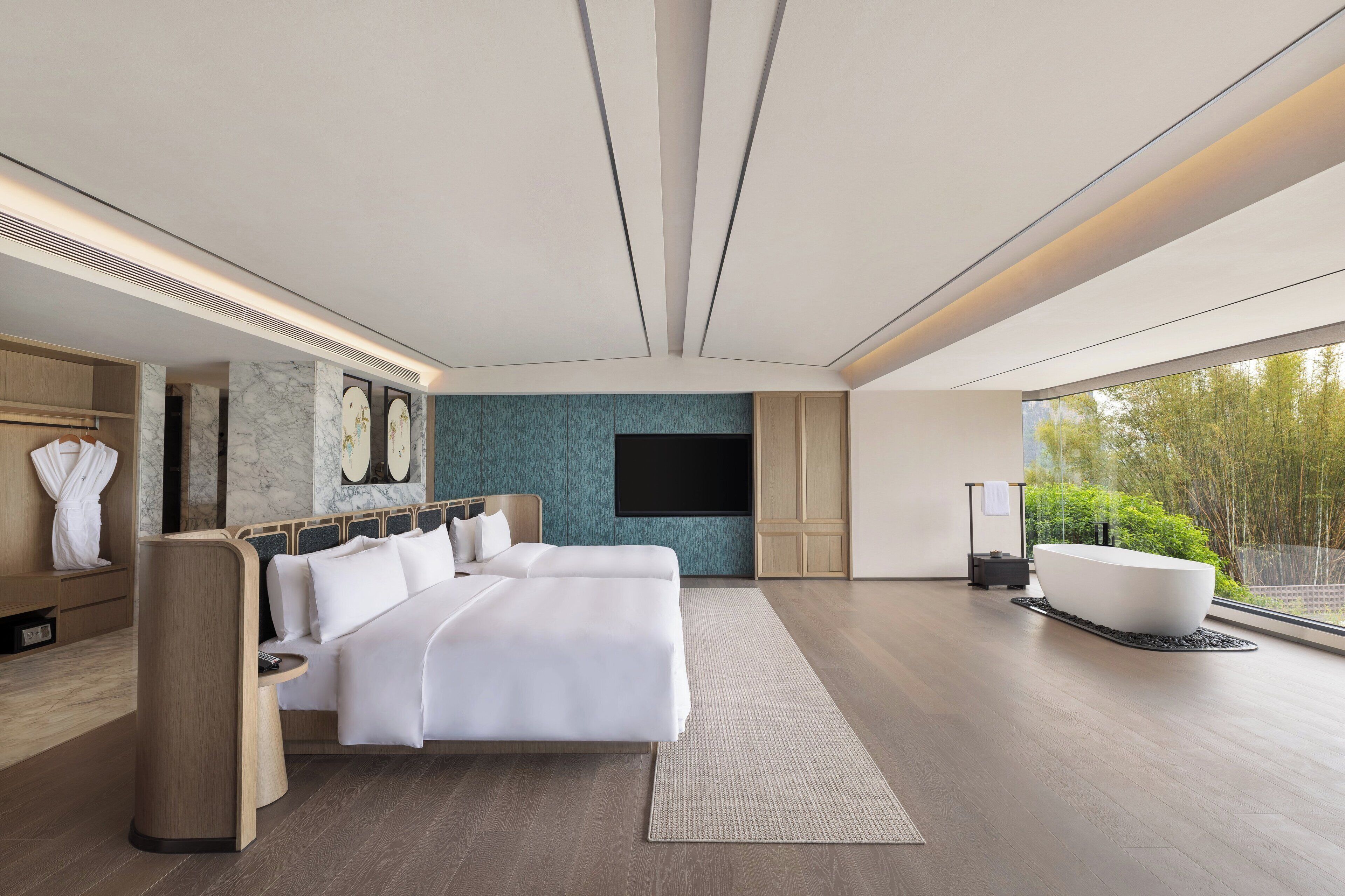A modern hotel room with a large bed, a bathtub, and a large window offering a view of greenery.