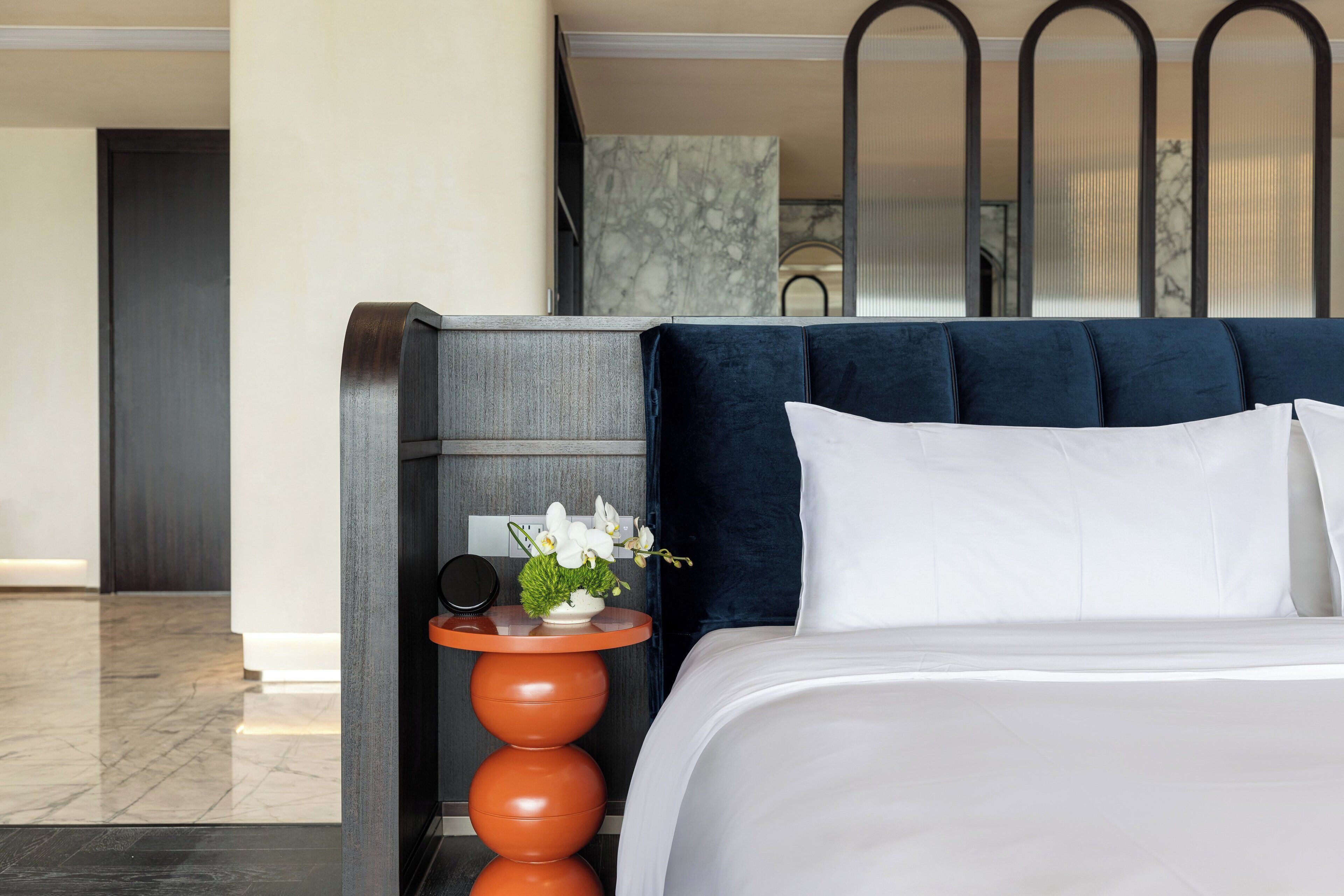 A hotel room with a bed, a nightstand with a vase of flowers, and a marble wall in the background.