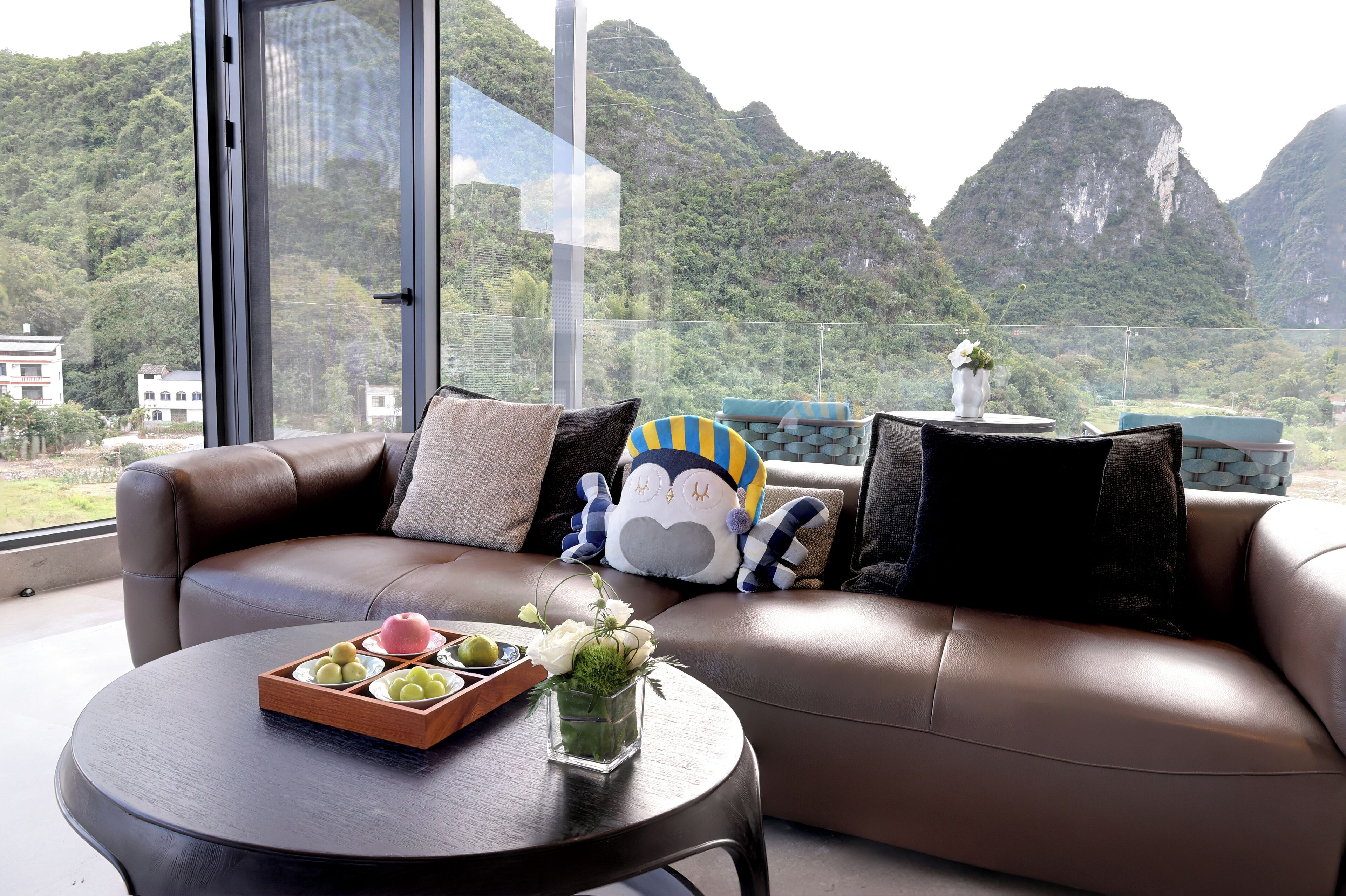 Una sala de estar moderna con un sofá de cuero, una mesa redonda de centro con una bandeja de bocadillos y un jarrón de flores. Un peluche está colocado sobre el sofá. La habitación tiene ventanas amplias que ofrecen vistas a montañas y vegetación.