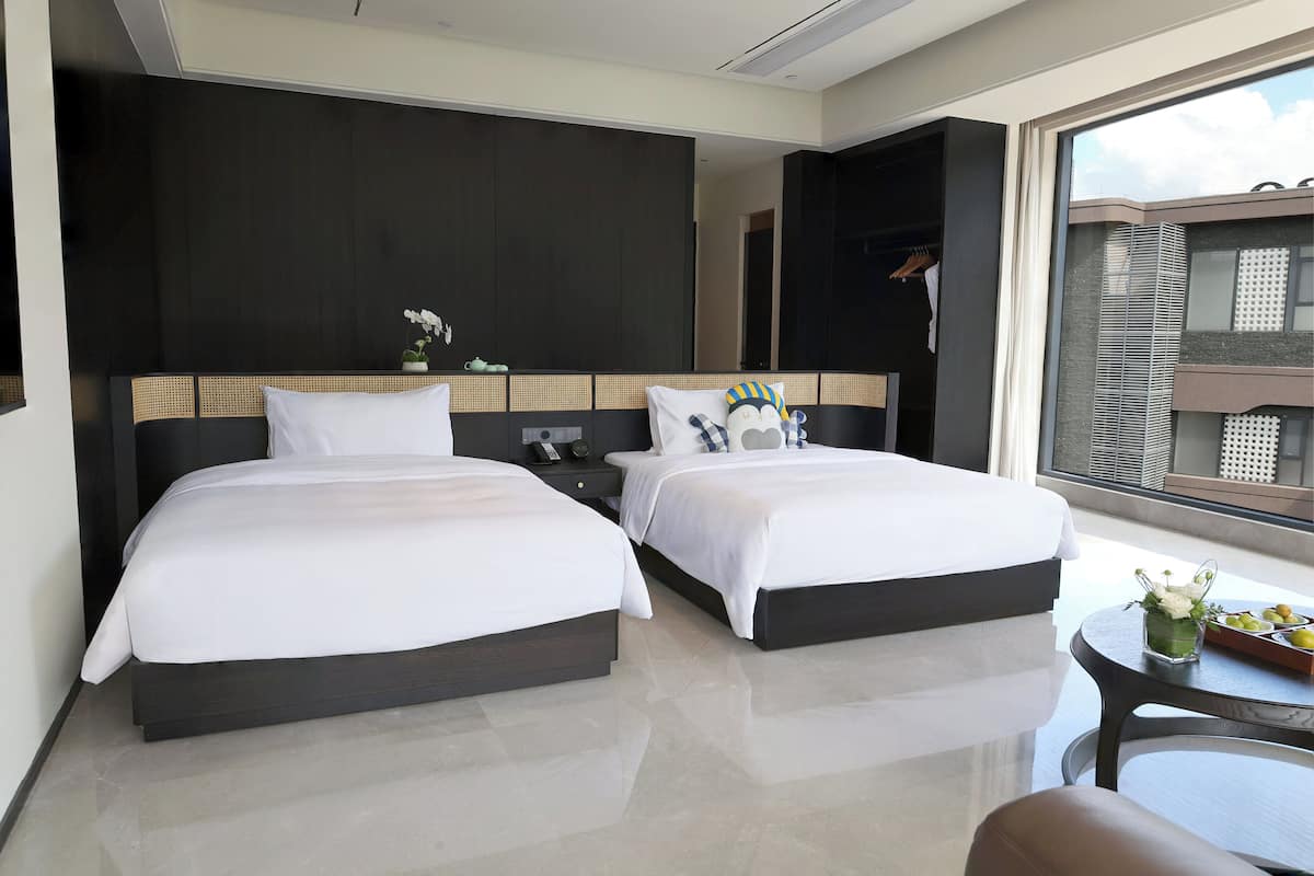 Habitación de hotel moderna con dos camas, una mesita de noche, una pequeña mesa con un florero y vistas al exterior.