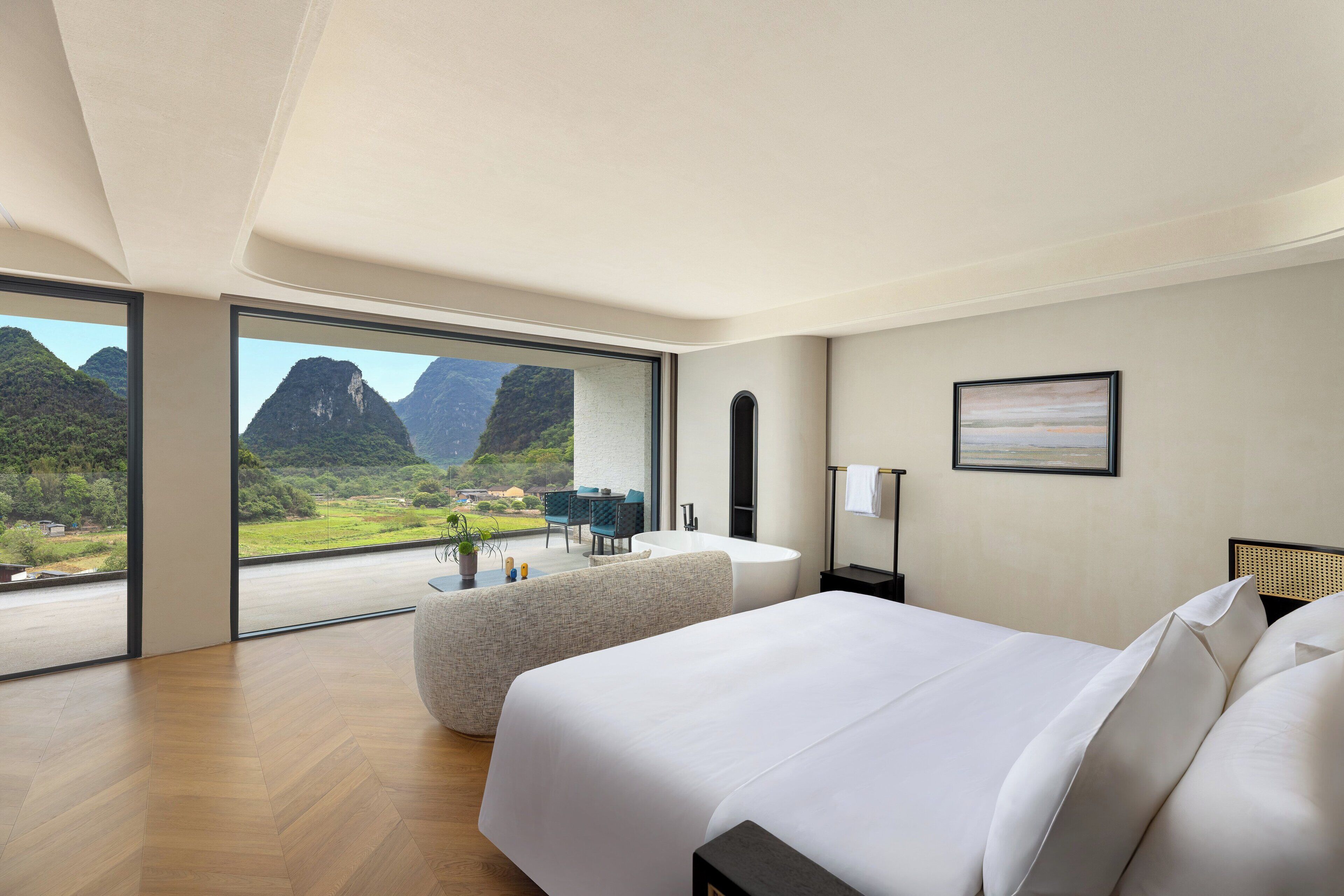 Habitación de hotel moderna con una cama grande, zona de estar con vistas a montañas y una pequeña mesa con una planta.