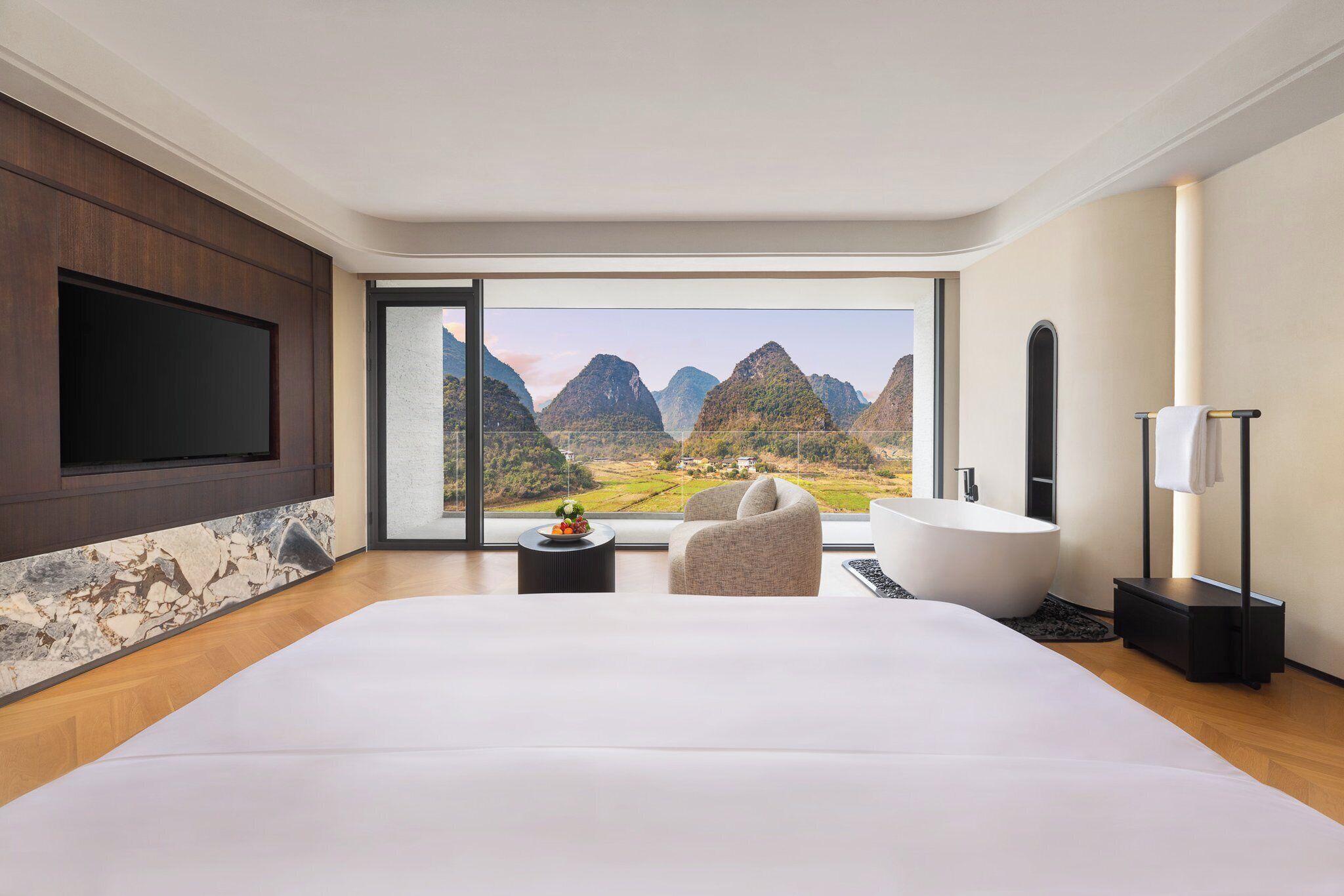 A modern hotel room with a large bed, a TV mounted on the wall, a bathtub, and a view of mountains through large windows.