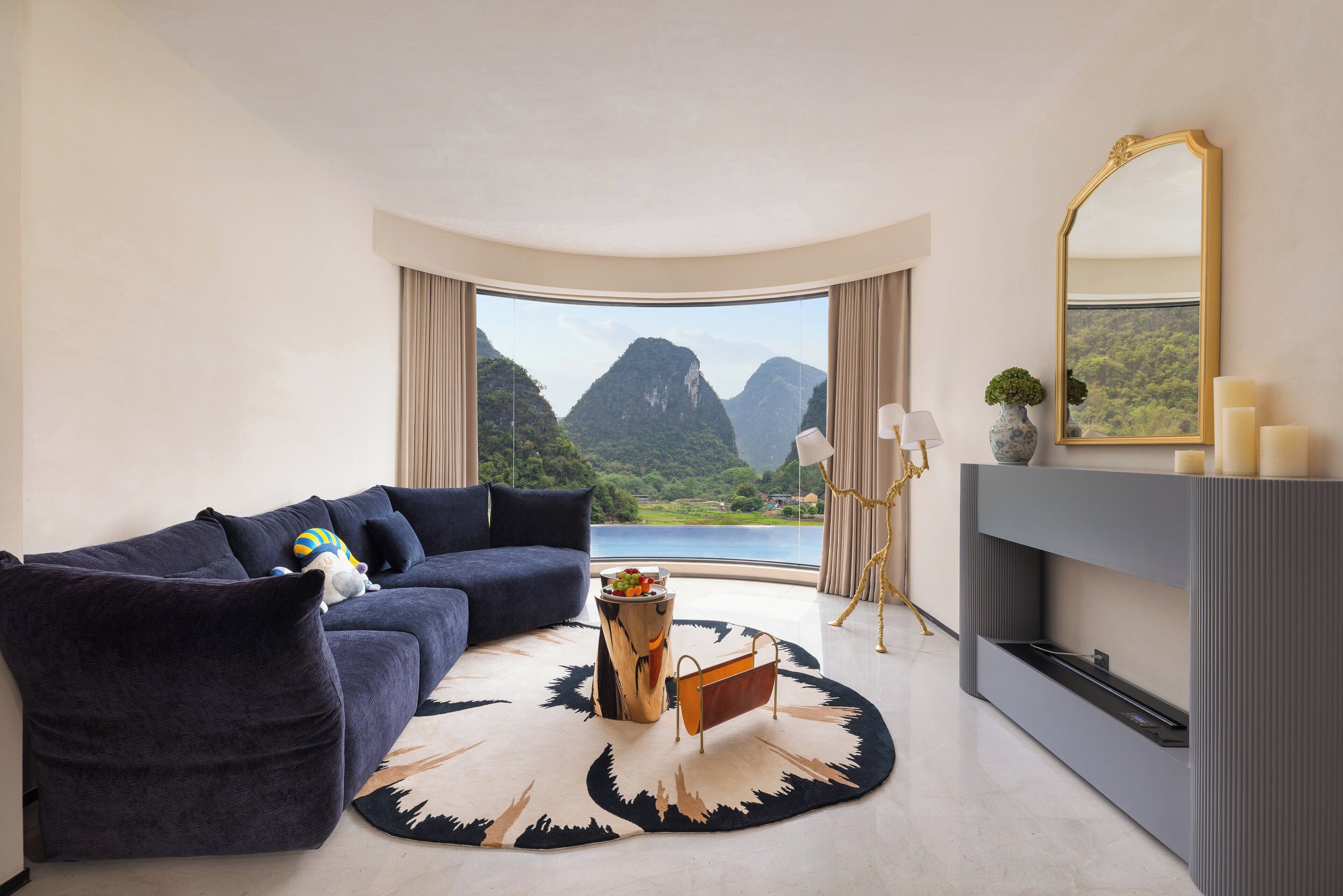 A modern living room with a curved sofa, a round rug, a fireplace, and a large window with a mountain view.