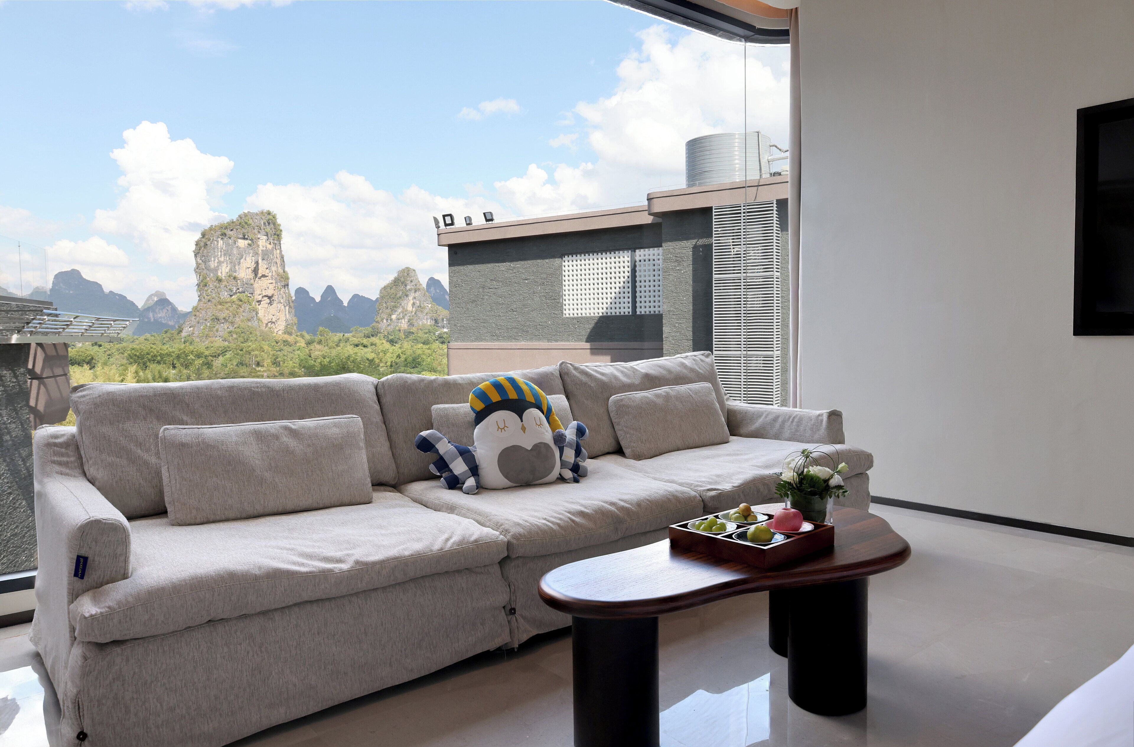Uma sala de estar moderna com um sofá, um brinquedo de pelúcia, uma mesinha com um tabuleiro e vista para montanhas através de grandes janelas.