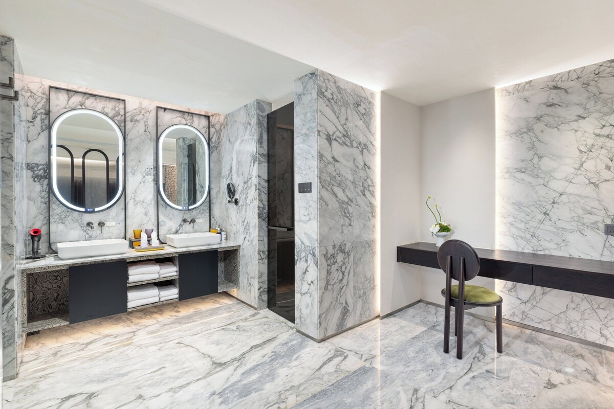 A modern bathroom with marble walls and floors, two sinks with mirrors, a towel rack, and a chair.