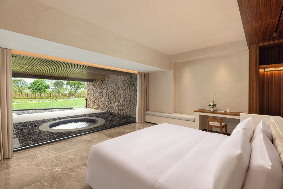 A modern hotel room with a large bed, a desk with a chair, and a view of a garden and outdoor area.