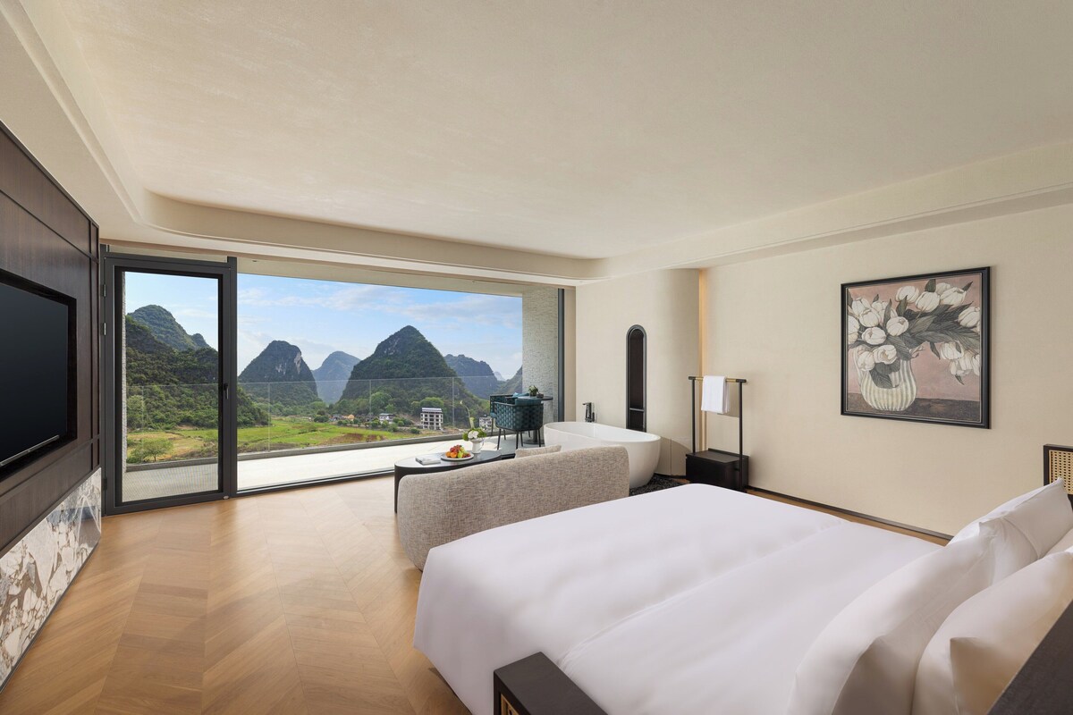 Une chambre d’hôtel avec un grand lit, un canapé, un bureau et une baignoire. Une grande fenêtre offre une vue sur les montagnes.