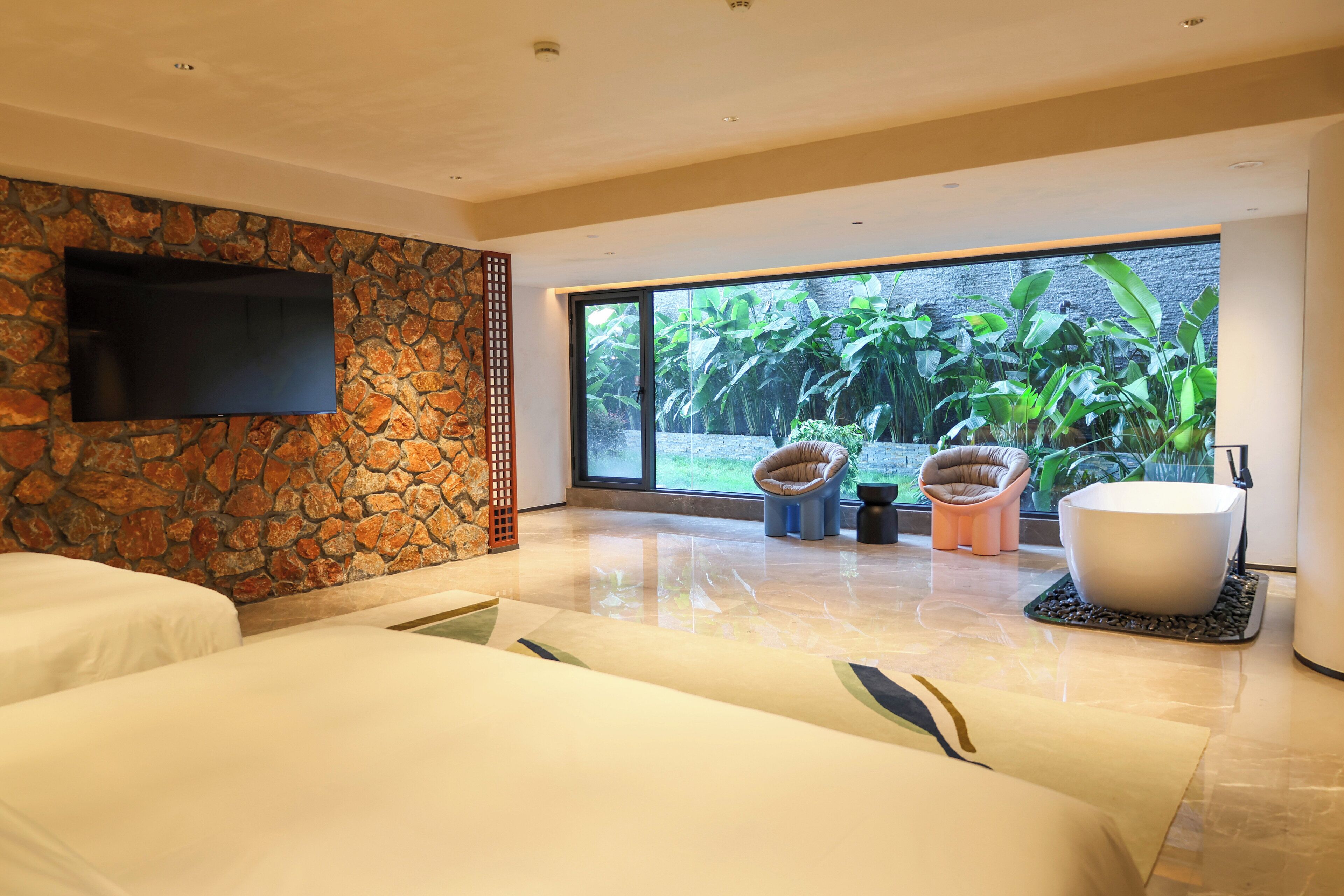 A modern hotel room with a stone wall, a flat-screen TV, a large window with a view of greenery, a bathtub, and two sofas.