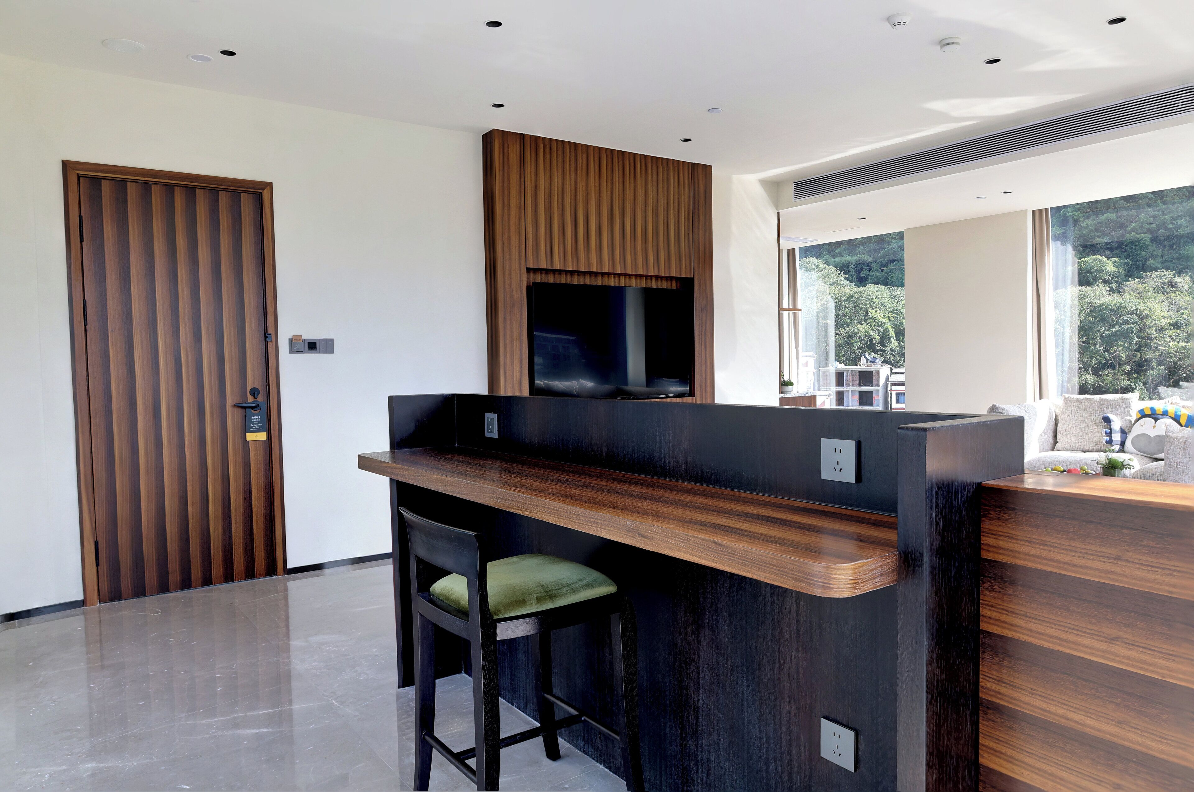 A modern reception desk with a wooden finish, a flat-screen TV mounted above it, and a view of a lush green landscape through large windows.
