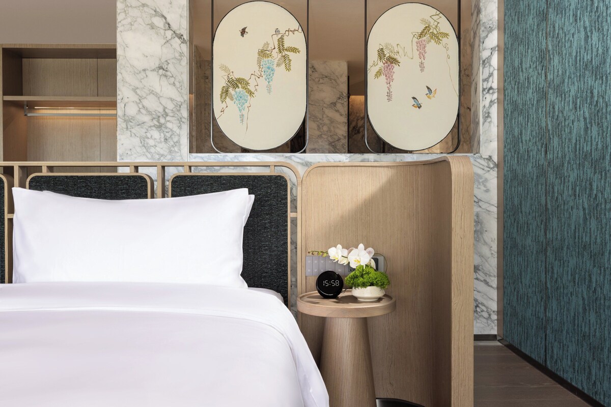 Une chambre d’hôtel comprenant un lit, une table de chevet avec une horloge et une plante, ainsi que des œuvres d’art décoratives accrochées au mur.