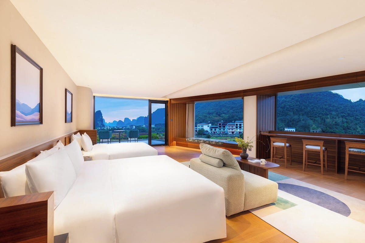 Una habitación de hotel moderna con dos camas grandes, una zona de estar, un escritorio y ventanas amplias con vistas a la montaña.