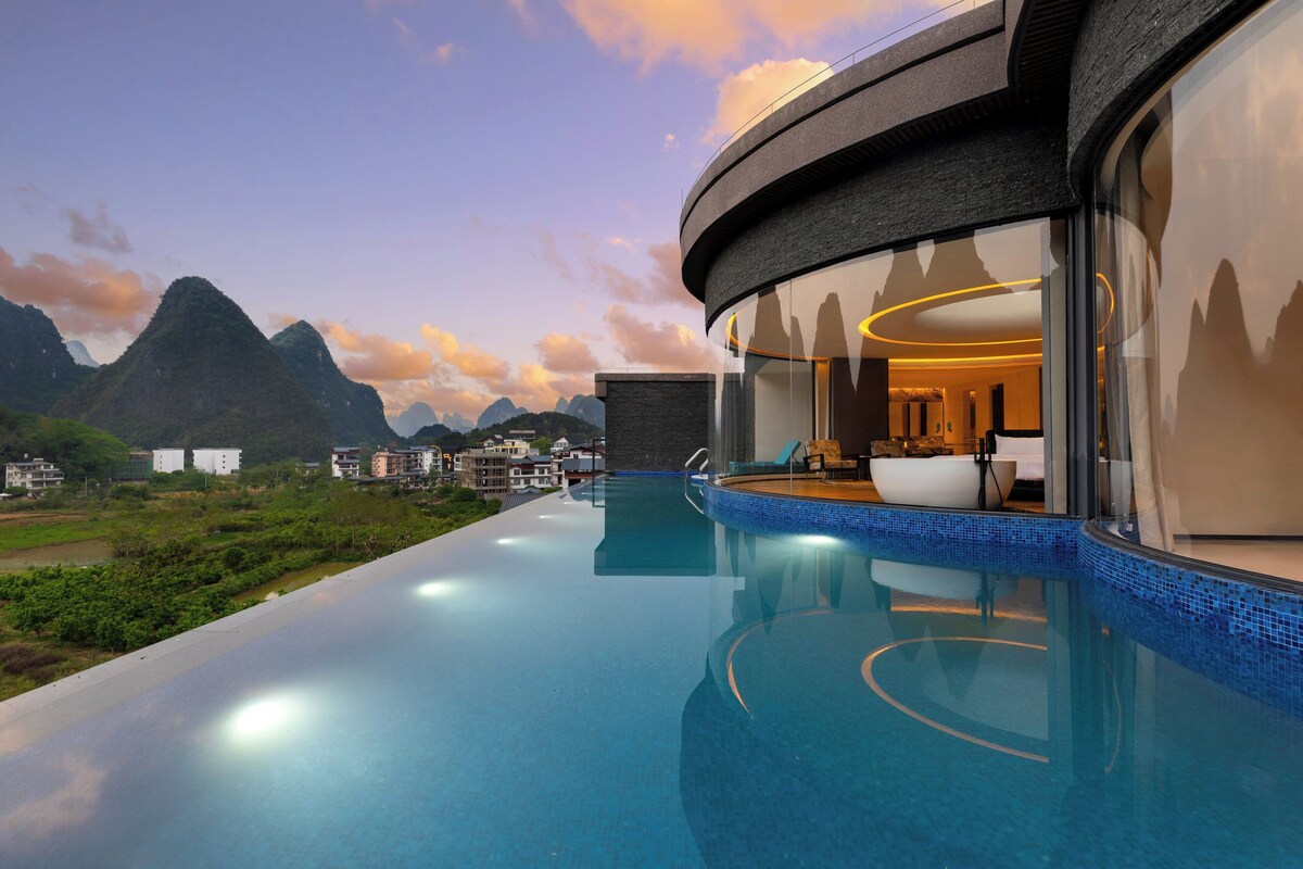 A modern infinity pool with a view of mountains, a well-lit interior, and a circular architectural design.