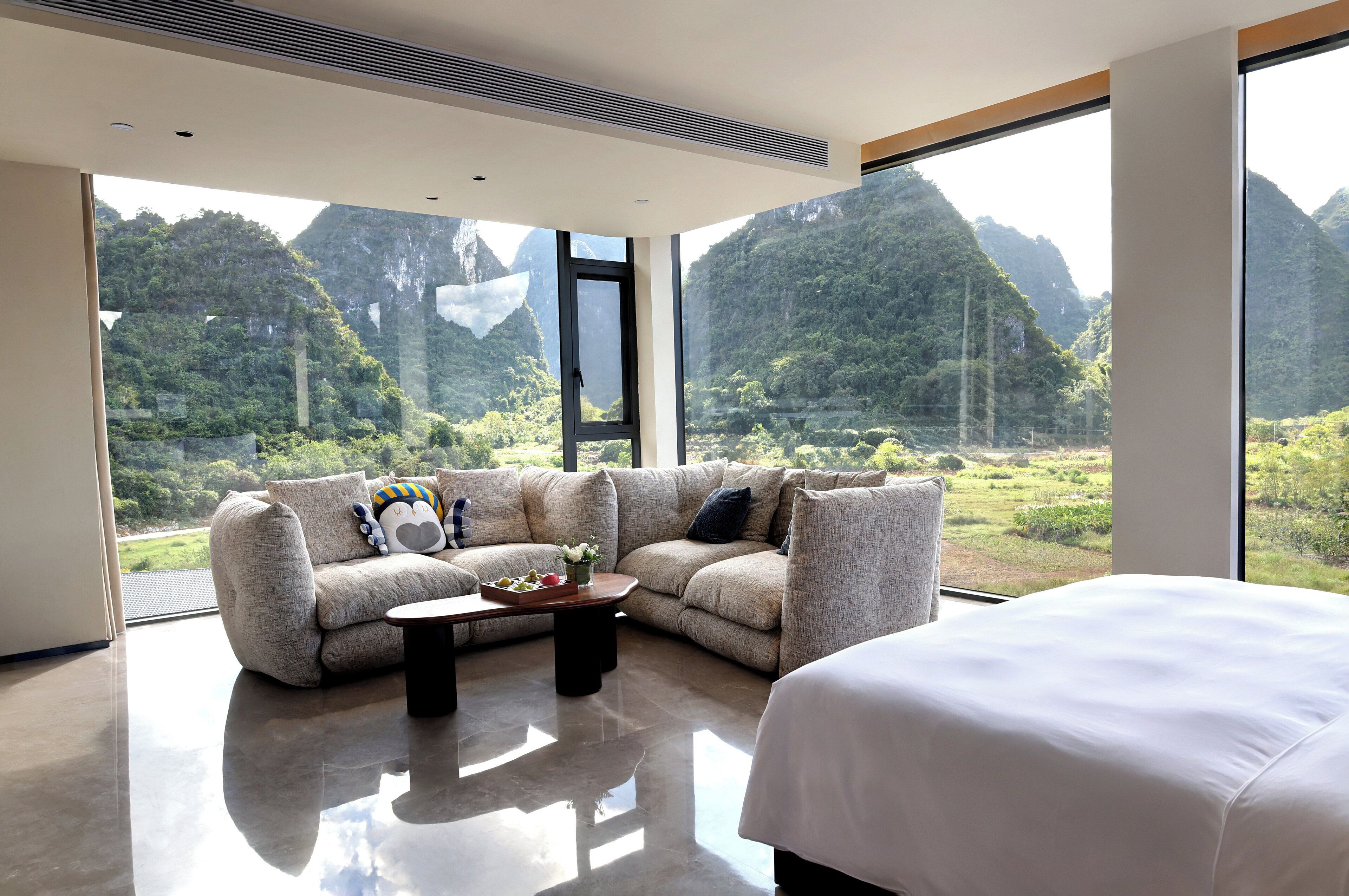 A modern room with a large bed, a sofa, and a coffee table. There is a large window offering a view of a mountainous landscape.