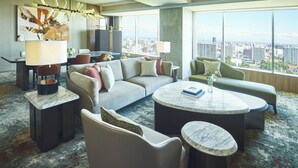 A modern hotel room with a large window offering a city view, a sofa, a round coffee table, and a dining area with a lamp.
