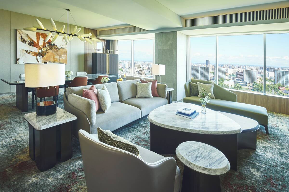 A modern hotel room with a large window offering a city view, a sofa, a round coffee table, and a dining area with a lamp.