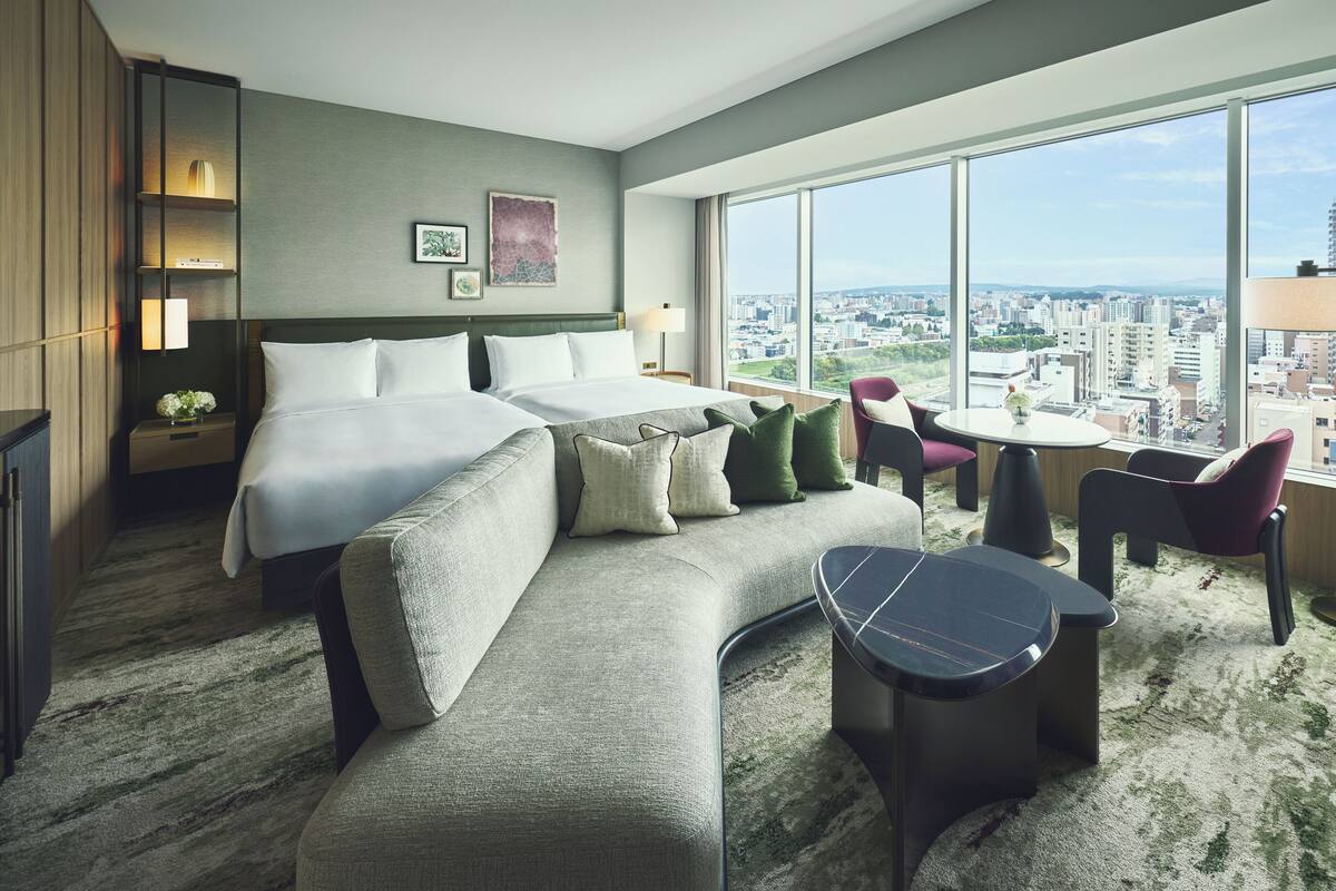 A modern hotel room with a large bed, a sofa, a small table, and a view of the city.