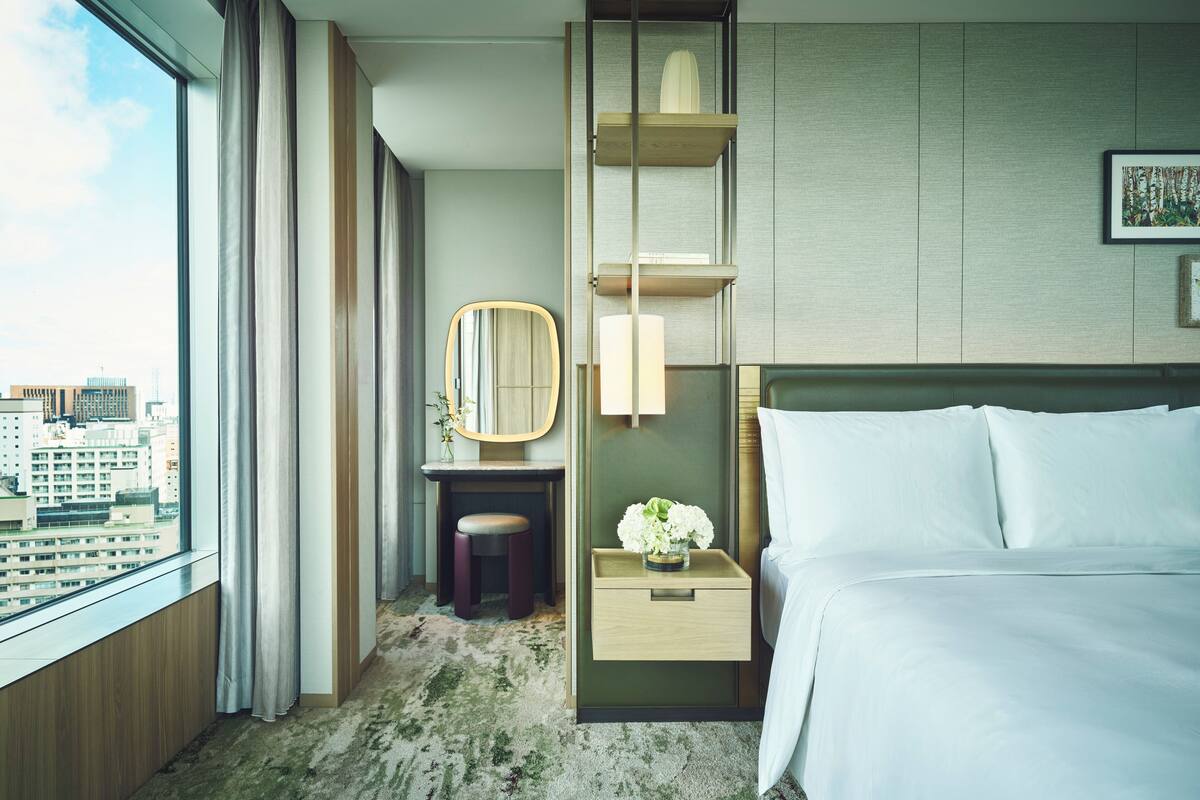 A hotel room with a bed, a desk, a chair, a mirror, a nightstand, and a view of the city.