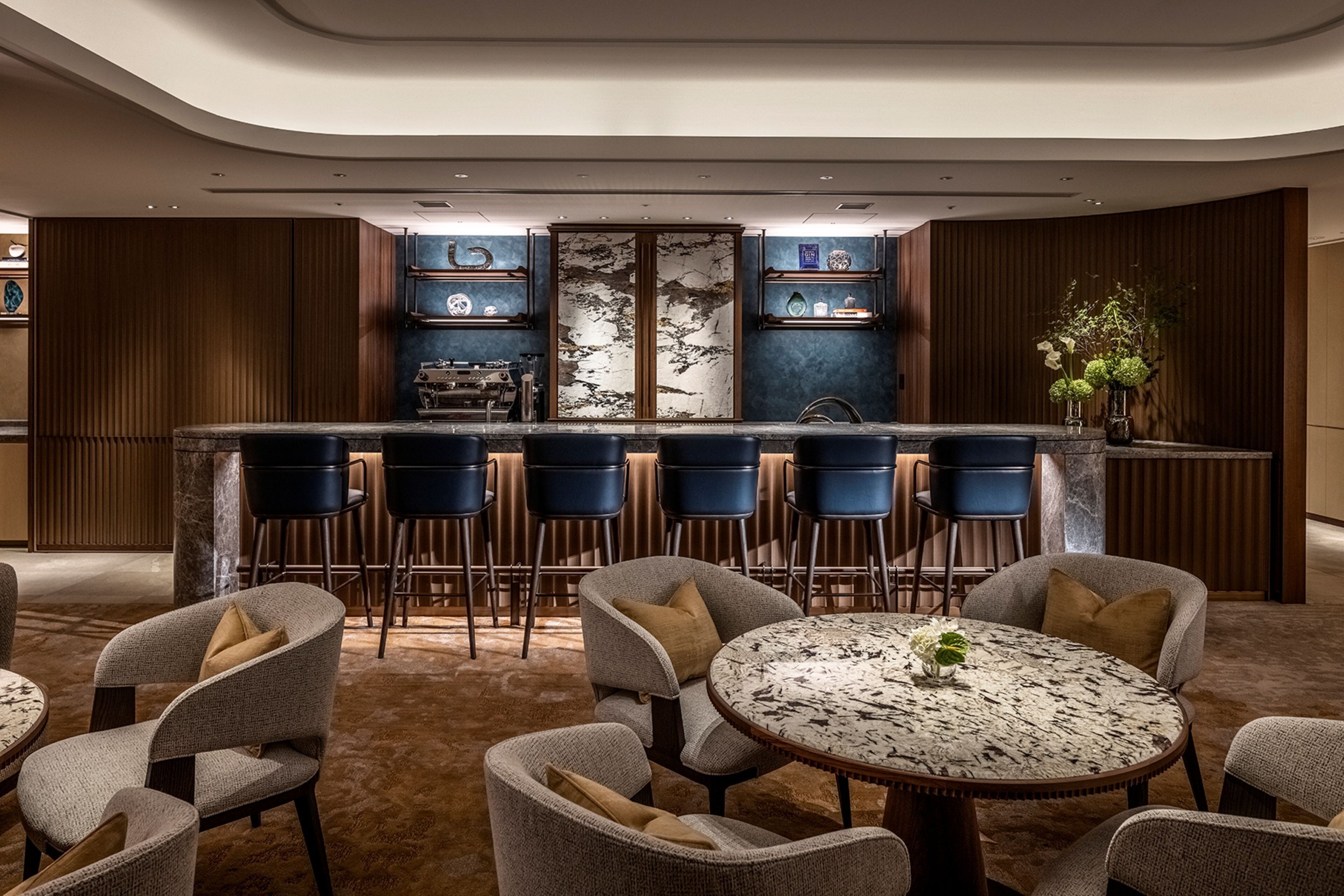 A modern bar area with a marble countertop, blue bar stools, and a round table with a vase of flowers.