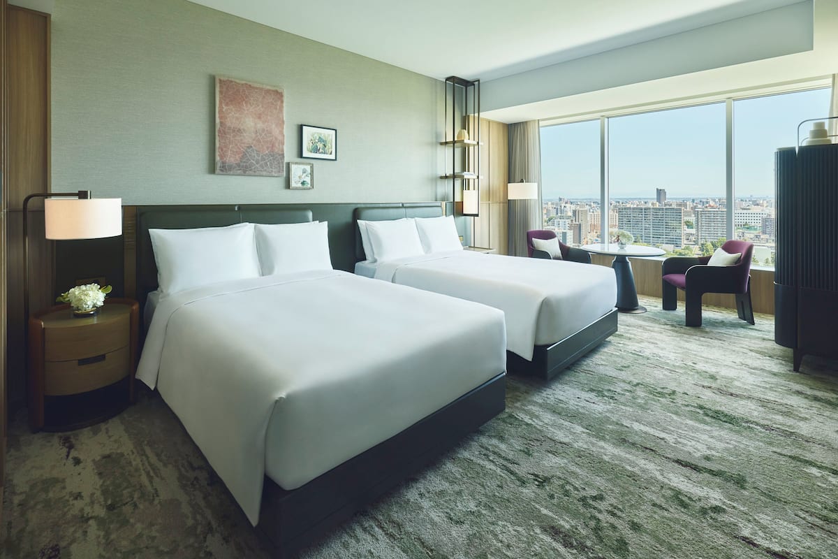 A hotel room with two beds, a desk, a chair, and a large window offering a city view.