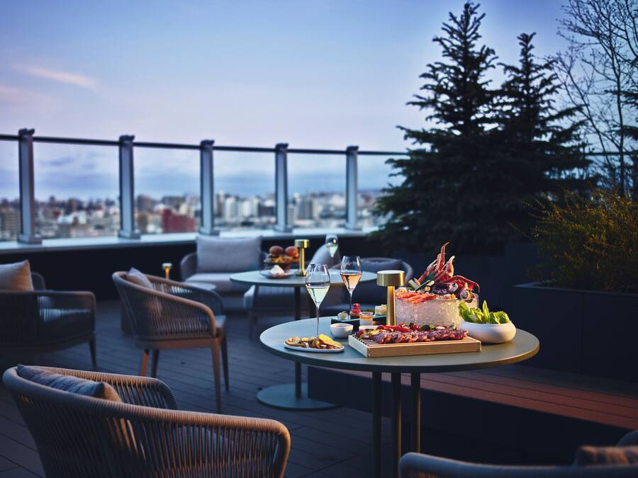 Eine Dachterrasse mit Außenmöbeln, einem Tisch mit Speisen und Getränken und Blick auf die Stadt.