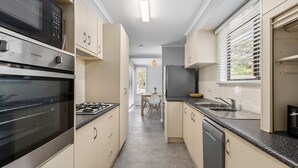 A compact kitchen with a gas stove, oven, and dishwasher, leading to a dining area with a table and chairs. - Bellarine Beach Shack (St Leonards)