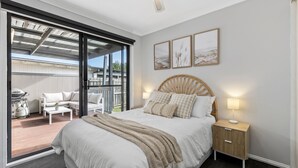 A bedroom with a large bed, a wooden nightstand, and a view of an outdoor patio with furniture. - Bellarine Beach Shack (St Leonards)