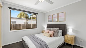 A bedroom with a large window, a bed with multiple pillows, a headboard, bedside tables, and wall art. - Bellarine Beach Shack (St Leonards)